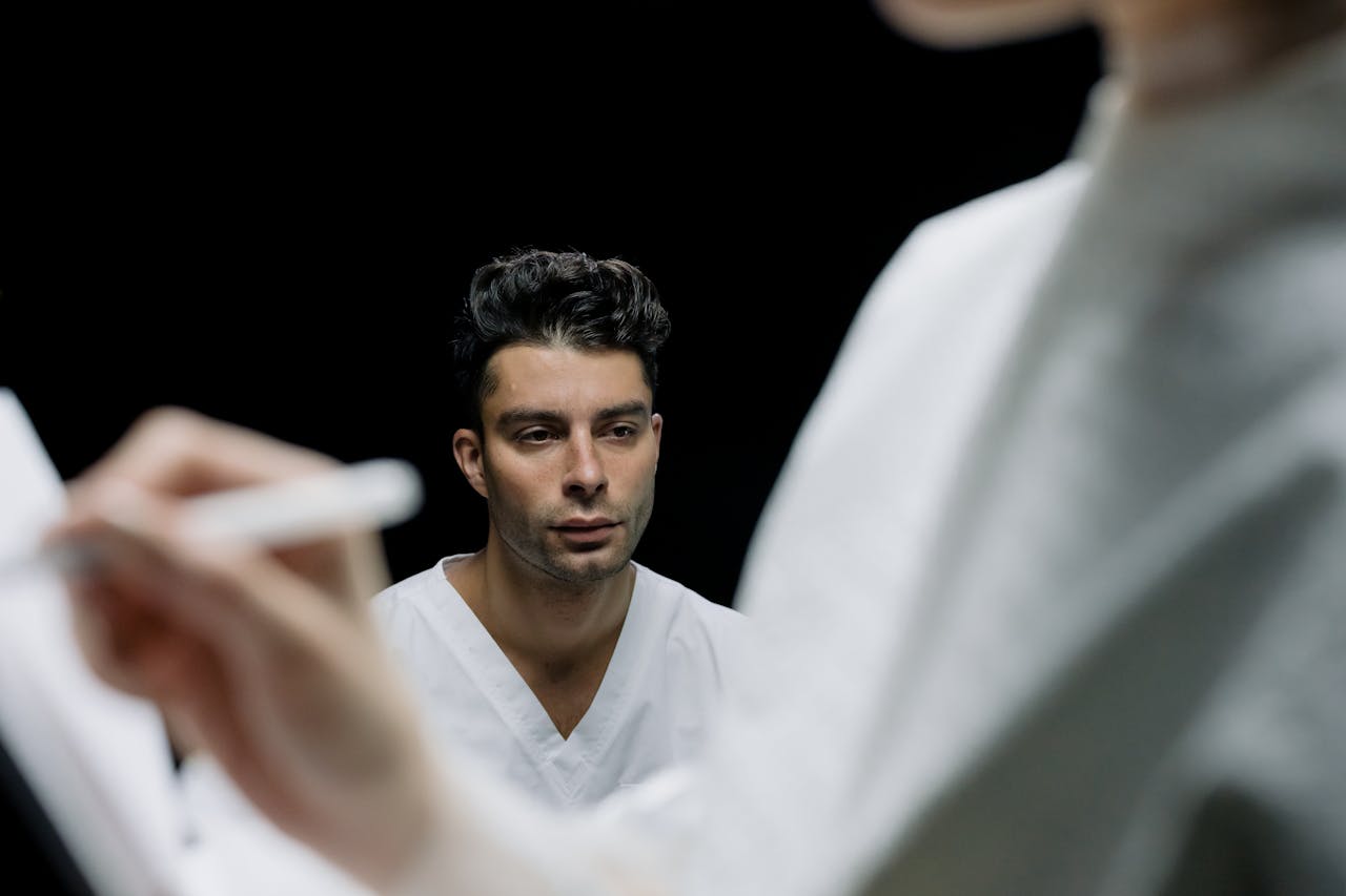 our-story An adult male with a serious expression in a medical environment, observed by a professional taking notes.